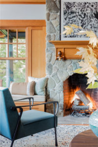 lakehouse,transitional living room furniture,picture windows,relaxed living room,stained moulding,stone fireplace surround,upholstered window bench cushion,velvet side chair,warm wood,window bench,wood beams,wood flooring,wood panel wall,wood paneling