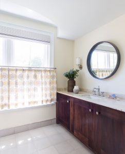 transitional bathroom design, café curtain in bathroom, round vanity mirror, custom bathroom millwork, white bathroom floor tile, dark wood bathroom vanity, beach house bathroom