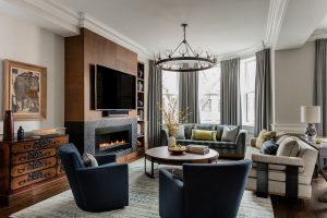 round coffee table, round cocktail table, fireplace, modern fireplace, traditional chest artwork, swivel chairs, bay windows, floor to ceiling drapery, modern sofa, round chandelier, metal and glass chandelier, glass table lamp, wall tv, living room, area rug